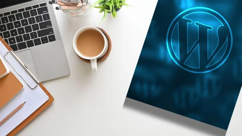 White table with a laptop, notebook, a mug with coffee and a dark blue panel with the wordpress logo.