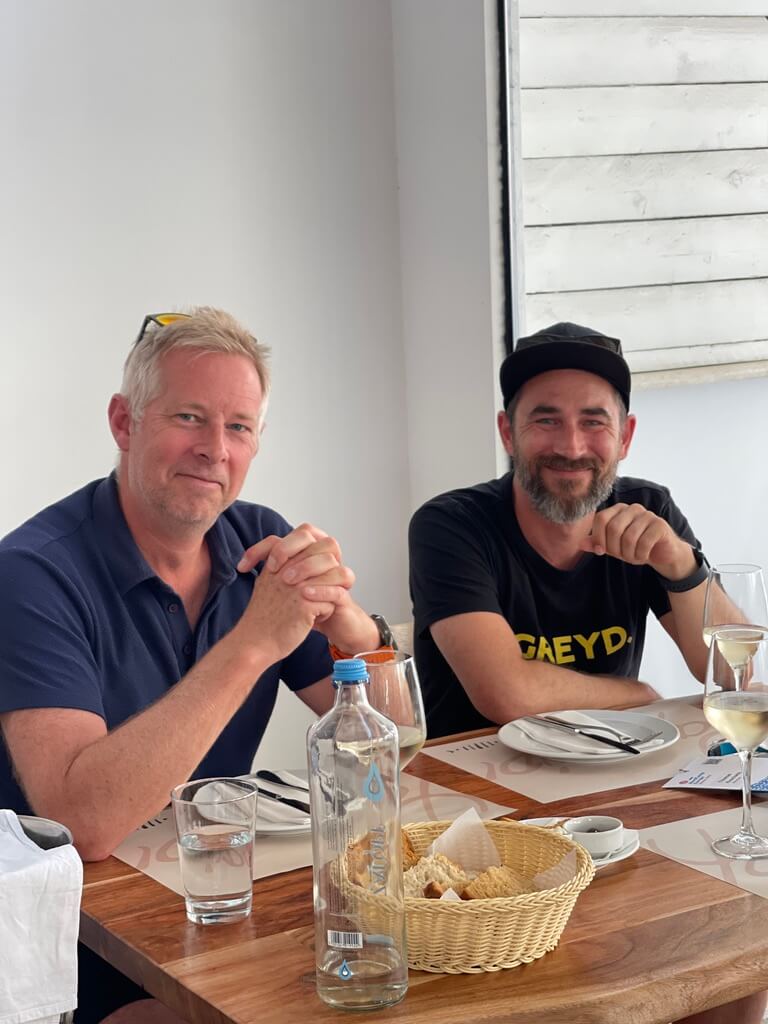 Two men sitting at a wooden table in a well-lit restaurant. The man on the left with short white hair, wearing a navy blue polo shirt and sunglasses resting on his head, is Jamie Marsland. He is sitting with his hands clasped together in front of him. The man on the right, Thomas Koschwitz, is wearing a black hat and a black t-shirt with the word “GREYD” visible on it. Both men are smiling.