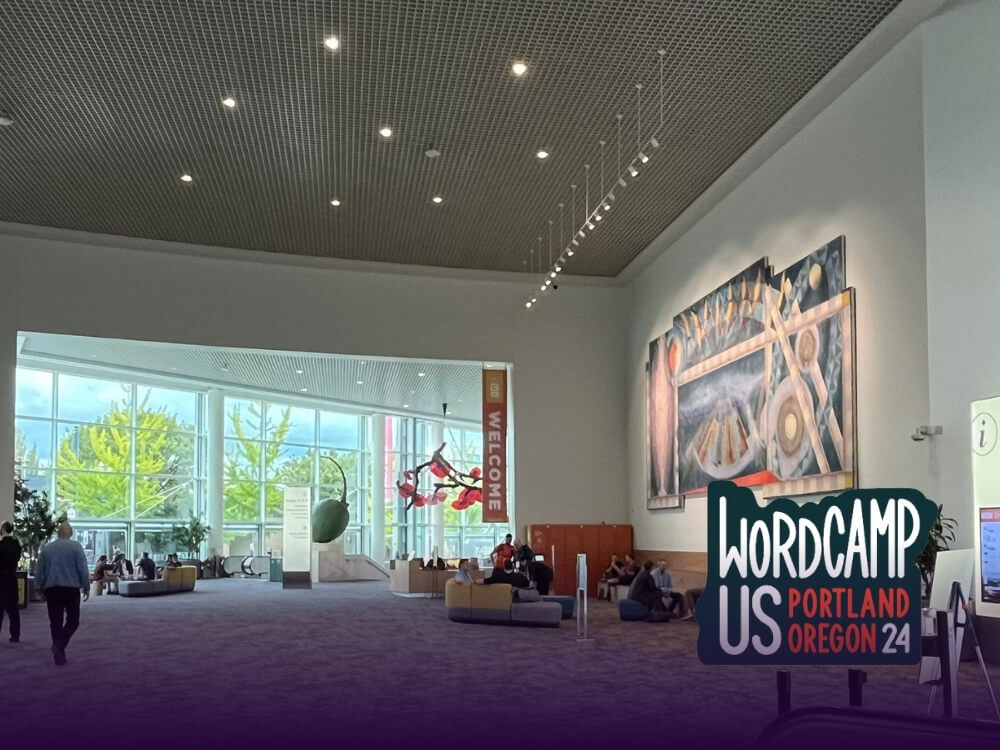 A spacious convention center lobby with large windows and artwork on the walls, featuring a ‘WordCamp US Portland Oregon 2024’ logo in the foreground.