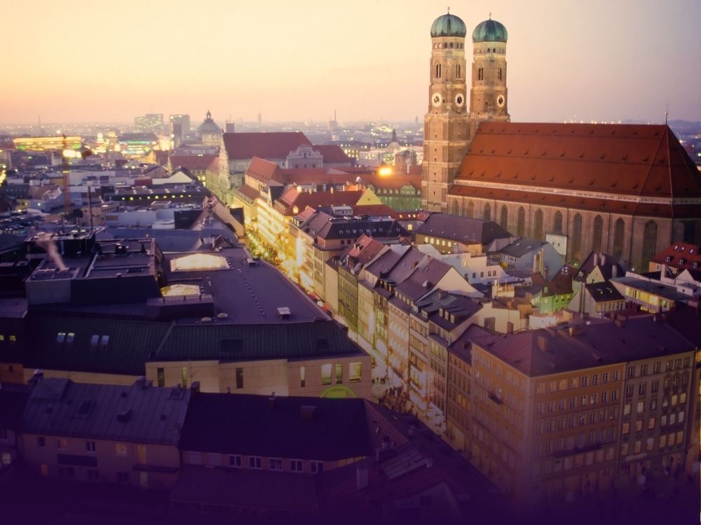 Abendansicht der Münchner Stadtlandschaft mit den beleuchteten Straßen und der ikonischen Frauenkirche mit ihren Zwillingstürmen unter einem sanften Sonnenuntergangshimmel.