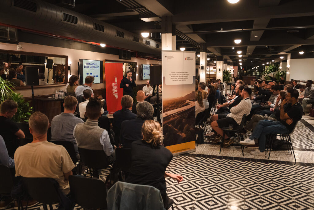 Audience attending the German Accelerator Startup Pitch Night, with Mark Weisbrod presenting in front of banners and screens displaying program visuals in a modern event space.