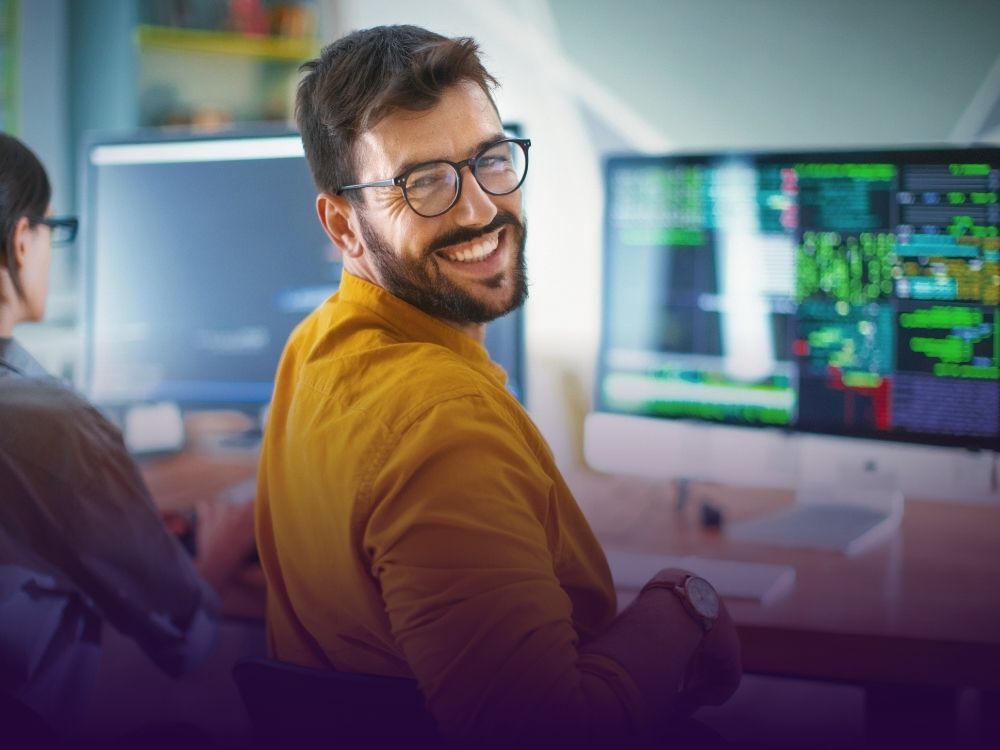 Ein lächelnder Mann mit Brille und Bart sitzt vor einem Computerbildschirm, auf dem Code zu sehen ist. Er trägt ein senfgelbes Hemd und schaut zur Kamera zurück.